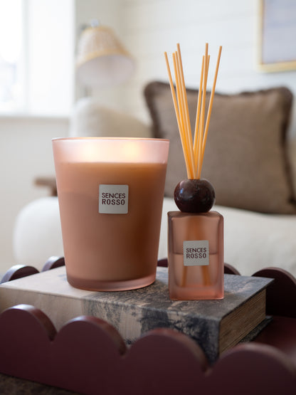 A Sences Rosso scented candle in a frosted glass jar and a matching reed diffuser bottle with wooden reeds, styled on a book in a softly blurred living room setting.