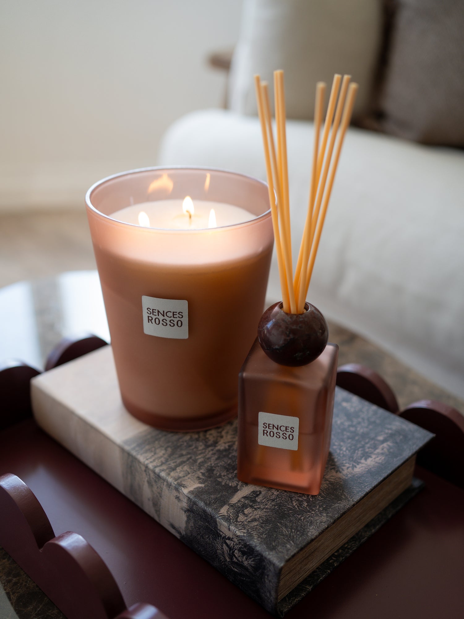 A lit Senses of Rosso scented candle in a frosted glass jar sits beside a matching reed diffuser bottle with wooden reeds on a stacked book in a softly blurred indoor setting.