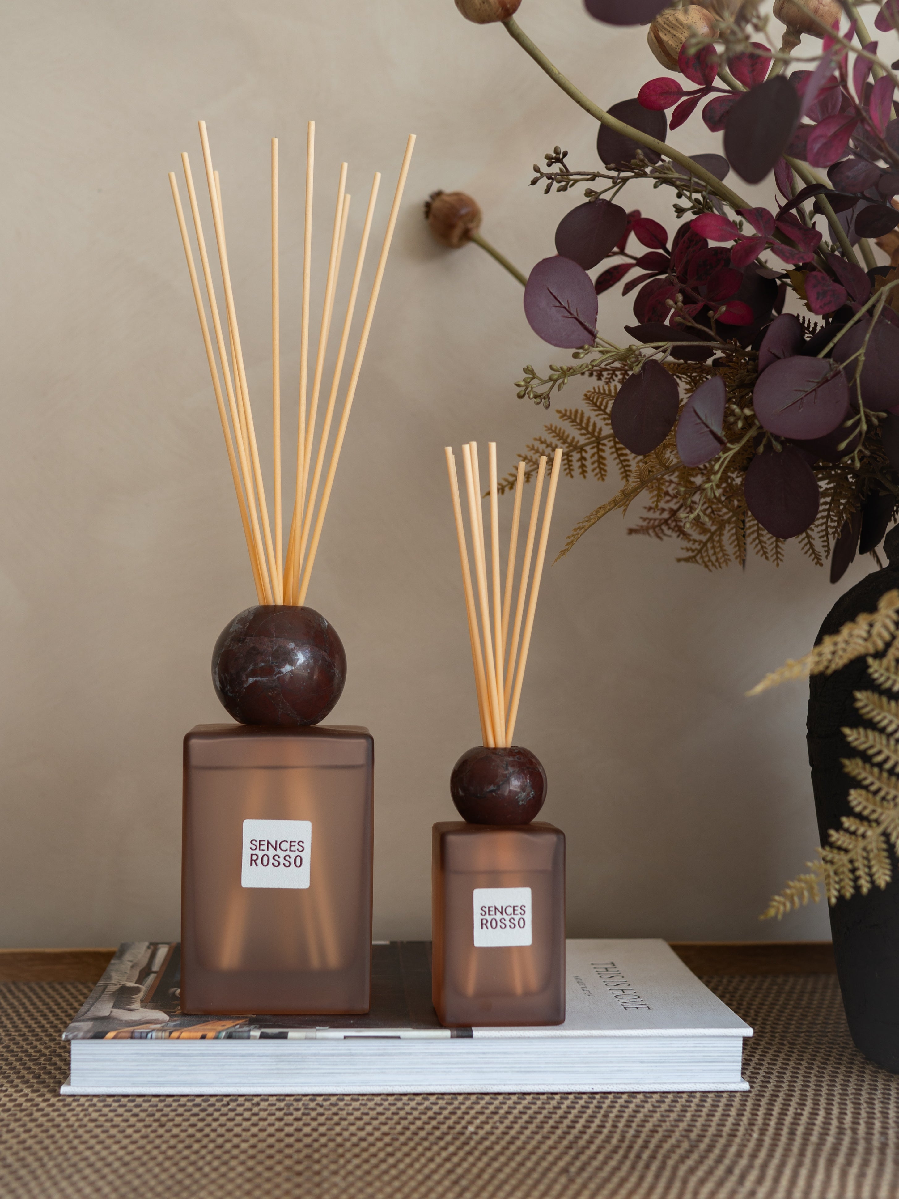 Two amber glass reed diffusers with dark round tops and natural reed sticks, displayed on a stack of books with dried floral decor in the background.