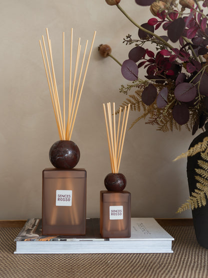 Two amber glass reed diffusers with dark round tops and natural reed sticks, displayed on a stack of books with dried floral decor in the background.