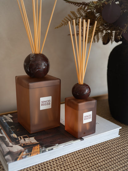 Two amber-brown reed diffusers with dark round tops and natural reed sticks, labeled &