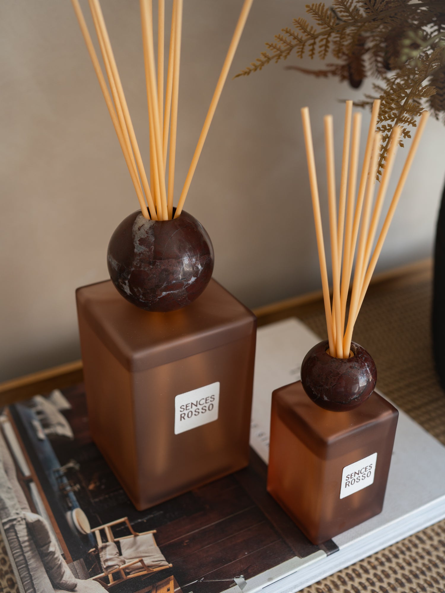 Two amber glass reed diffusers with dark round stone tops and natural reed sticks, displayed on a tabletop with a magazine and dried foliage in the background. The larger diffuser has a visible label reading SENCES ROSSO.