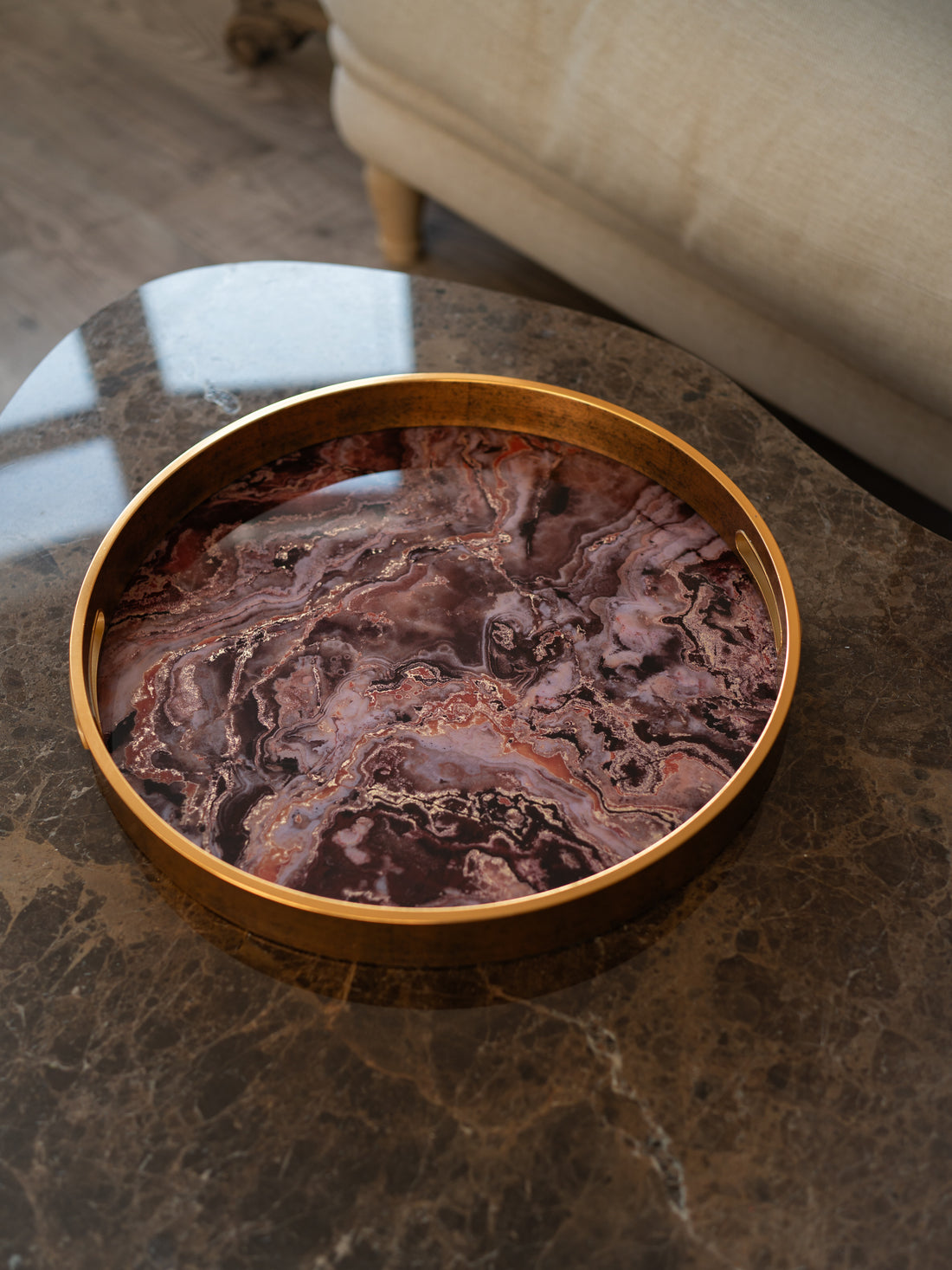 A round gold-framed tray with a polished reddish-brown and purple agate pattern, resting on a dark marble table in a modern living room setting.
