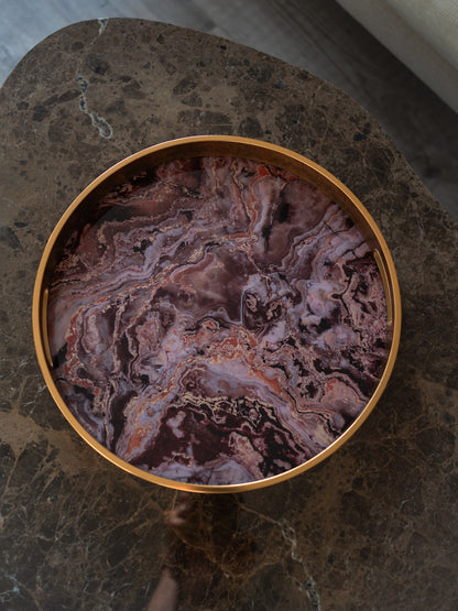 A round decorative tray with a polished gold-toned rim and a marbled purple, pink, and brown agate-like pattern, shown from above on a dark stone surface.