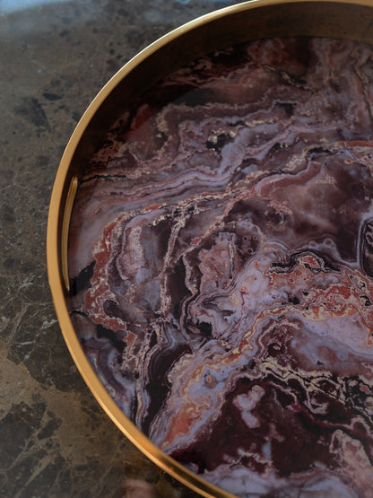 A close-up of a round decorative tray with a polished gold-toned rim and a marbled stone insert featuring swirling patterns of purple, burgundy, brown, and cream, set on a dark marble surface.