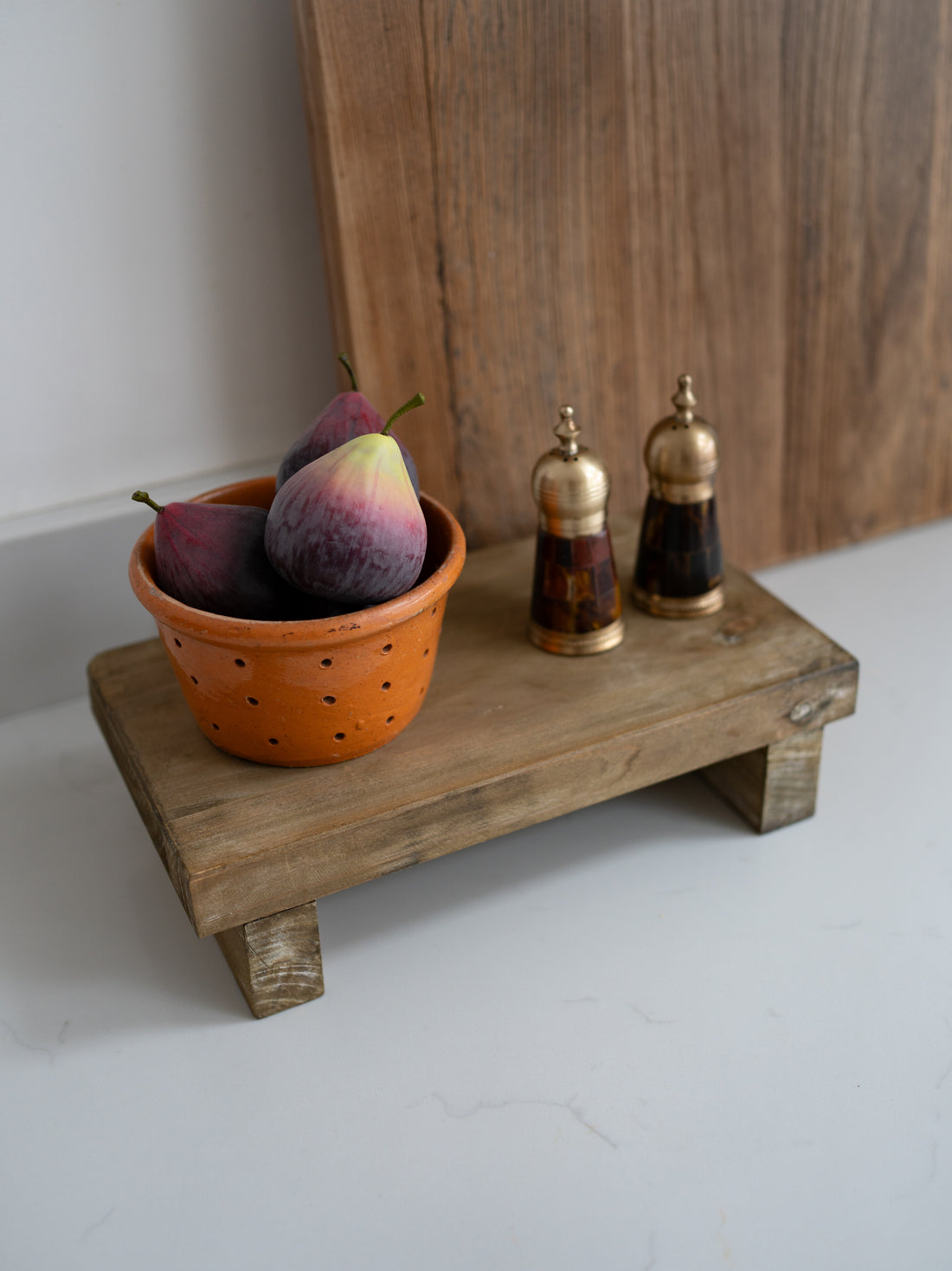 Small wooden riser with a colander of figs, two small bottles, and a wooden board in the background.