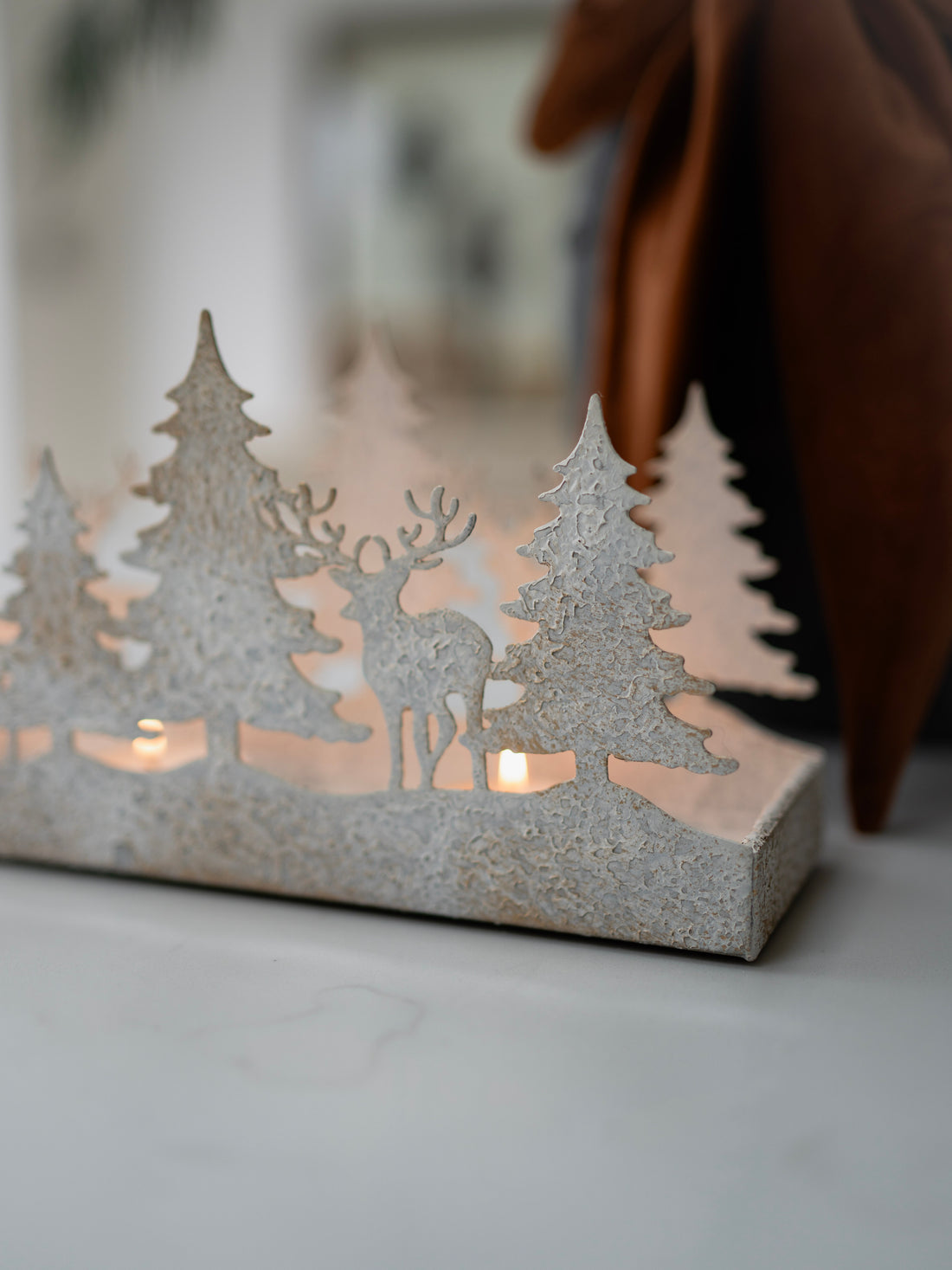 Decorative candle holder with cut-out trees and deer on a white surface.