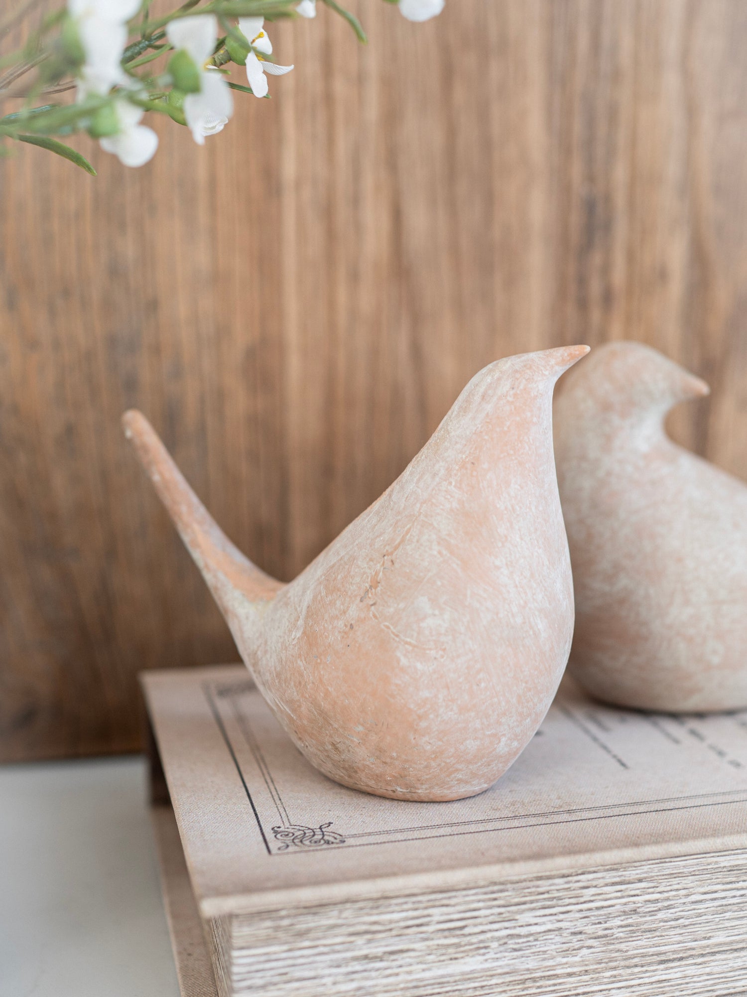 Two beige bird-shaped sculptures on a wooden surface with a blurred floral background