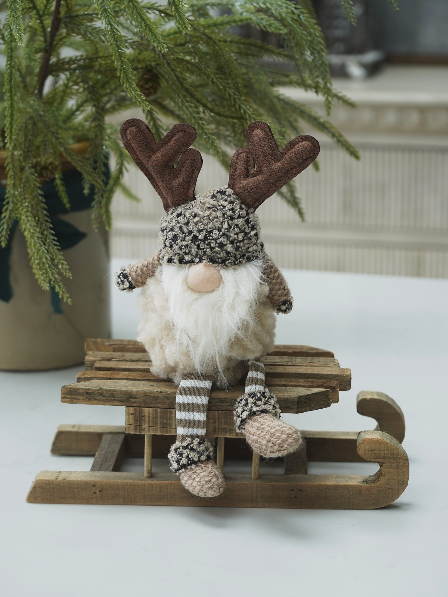 Decorative gnome with reindeer antlers on a small wooden sled, sitting on a white surface with a plant in the background.