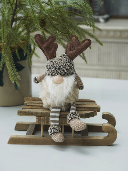 Decorative gnome with reindeer antlers on a small wooden sled, sitting on a white surface with a plant in the background.