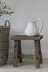 A rustic wooden stool made from pine wood, displayed in a room setting with a white ceramic jug on top.