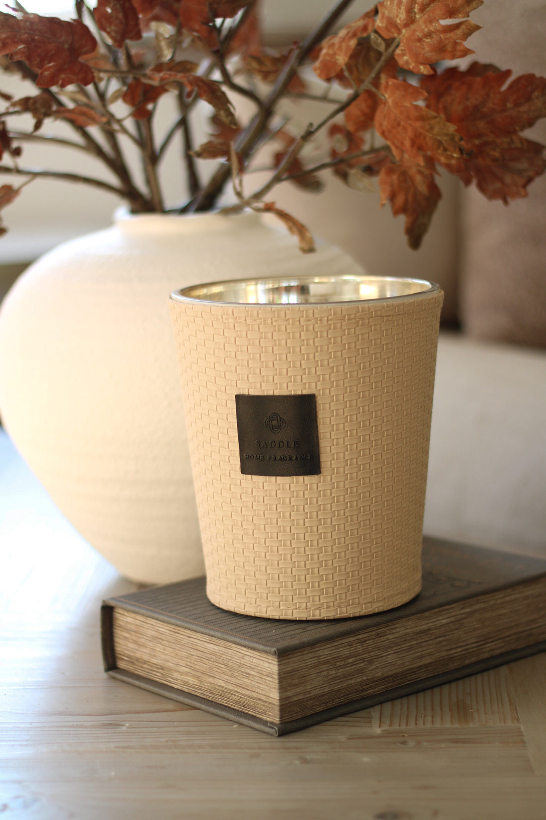 Beige textured candle with a black label on a wooden surface, with a blurred background of a vase and leaves.