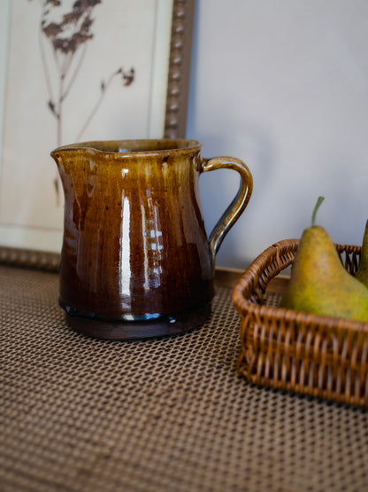 Brown ceramic pitcher on a textured surface with a basket of pears in the background.