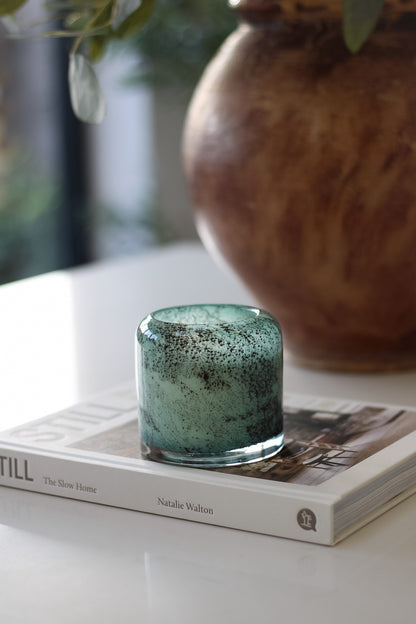 Teal glass candle holder on a book with a blurred vase in the background