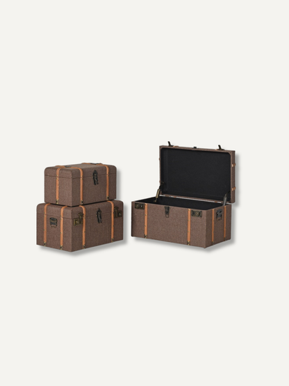 Three vintage-style storage trunks in a brown woven fabric with tan straps and black trim, shown on a light gray background. One trunk is open, revealing a black interior, while two others are closed and stacked.
