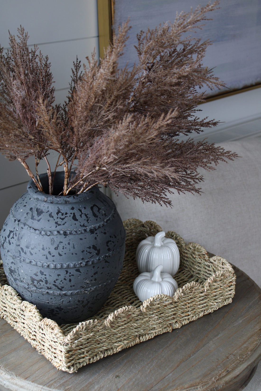 Decorative arrangement with a textured vase, dried plants, and a white pumpkin on a scalloped seagrass woven tray.