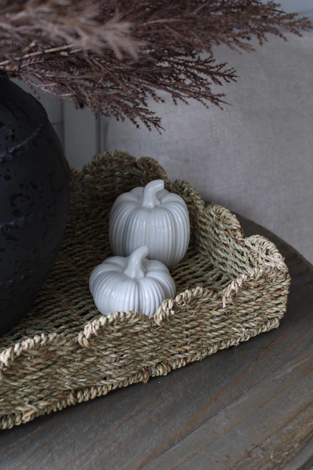 Two white pumpkins on a scalloped seagrass woven tray with decorative branches in the background.