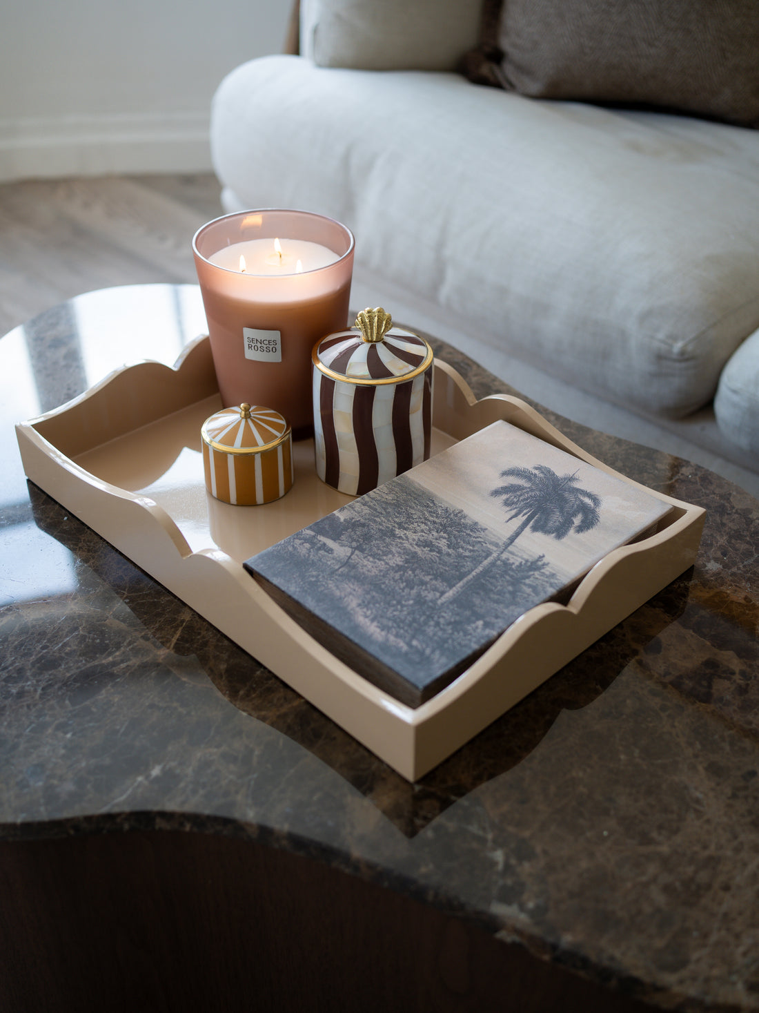 A styled home decor arrangement on a dark marble coffee table, featuring a beige scalloped tray holding a lit pink candle, striped jars, and a slate coaster with a palm tree print, with a light-colored sofa in the background.