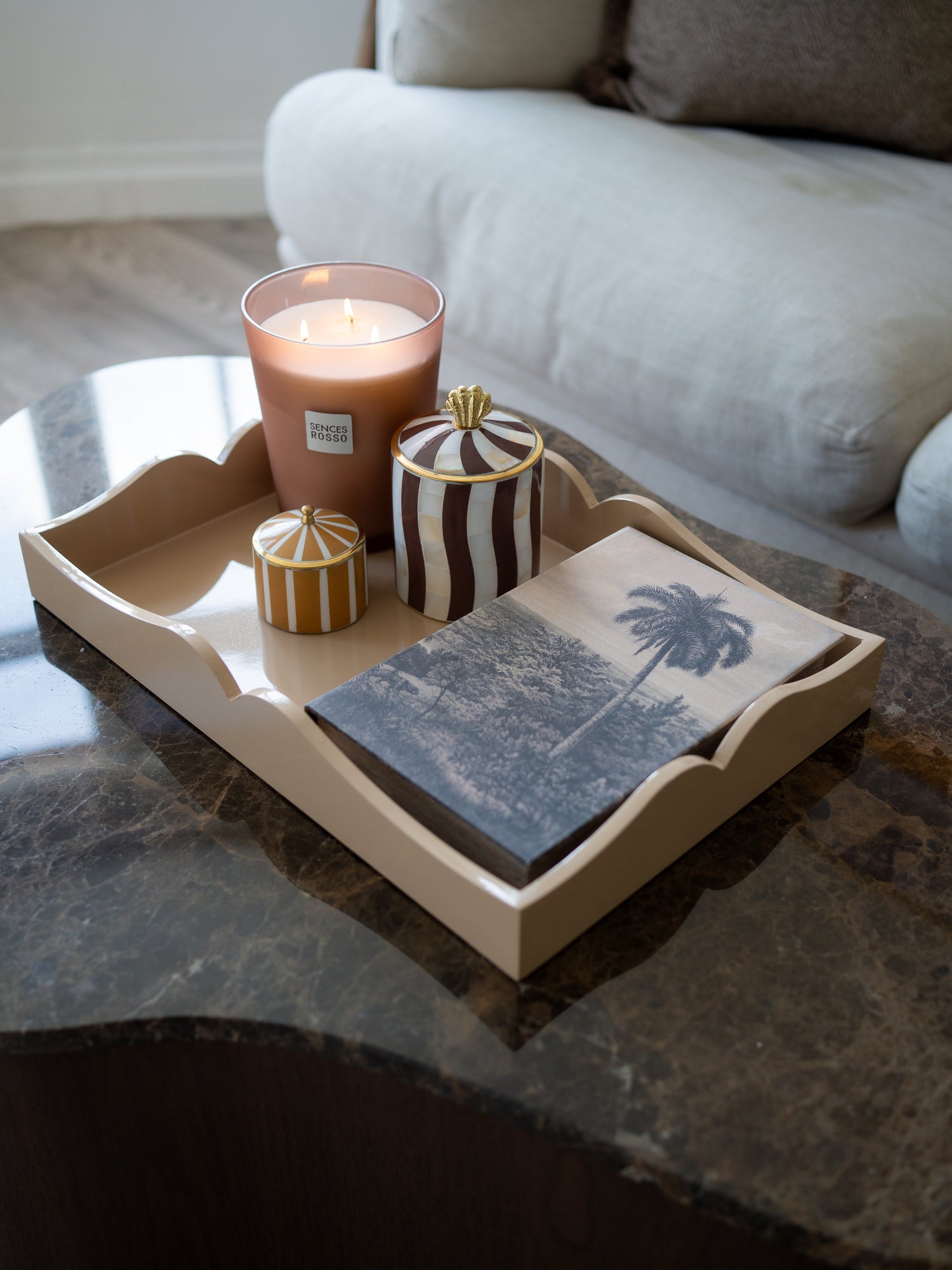 A styled home decor arrangement on a dark marble coffee table, featuring a beige scalloped tray holding a lit pink candle, striped jars, and a slate coaster with a palm tree print, with a light-colored sofa in the background.
