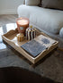 A styled home decor arrangement on a dark marble coffee table, featuring a beige scalloped tray holding a lit pink candle, striped jars, and a slate coaster with a palm tree print, with a light-colored sofa in the background.