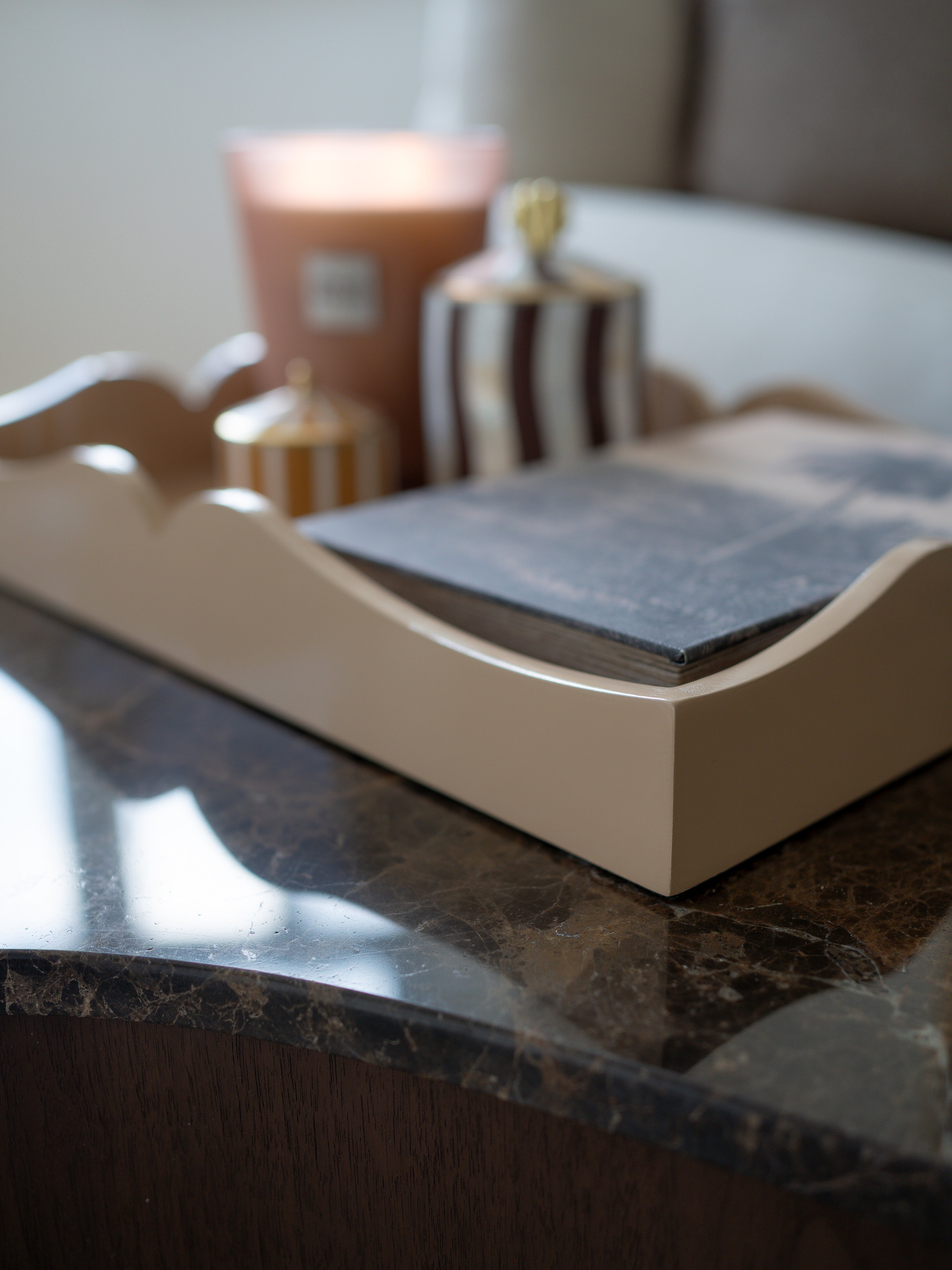 A close-up of a cream-colored wooden tray with scalloped edges, holding a dark slate notebook, styled on a dark marble table with candles in the background.
