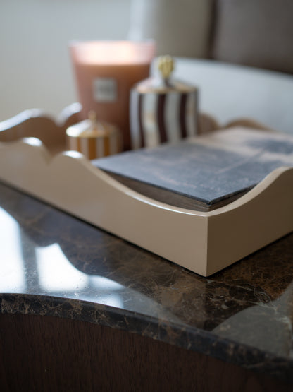 A close-up of a cream-colored wooden tray with scalloped edges, holding a dark slate notebook, styled on a dark marble table with candles in the background.