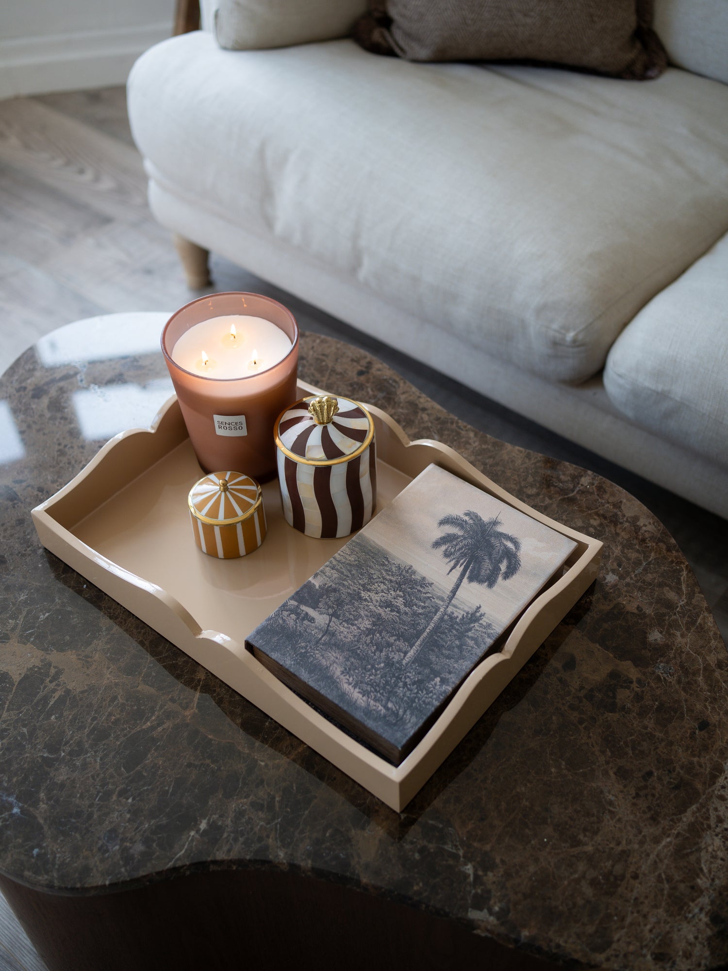 A styled home interior scene showing a beige wooden serving tray on a dark marble coffee table, holding a lit terracotta candle, two small decorative jars, and a folded napkin with a palm tree print. A light-colored sofa is visible in the background.