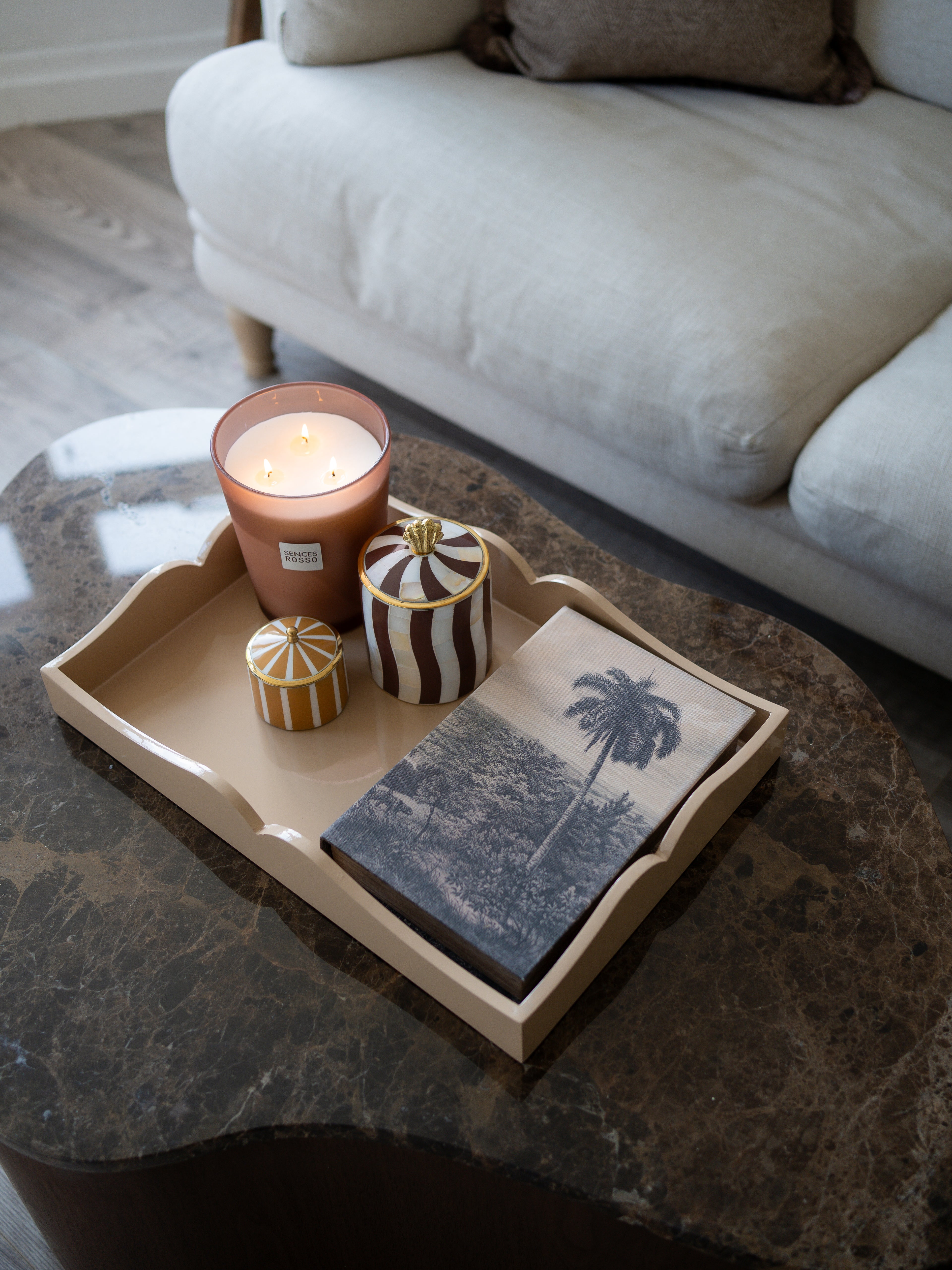 A styled home interior scene showing a beige wooden serving tray on a dark marble coffee table, holding a lit terracotta candle, two small decorative jars, and a folded napkin with a palm tree print. A light-colored sofa is visible in the background.