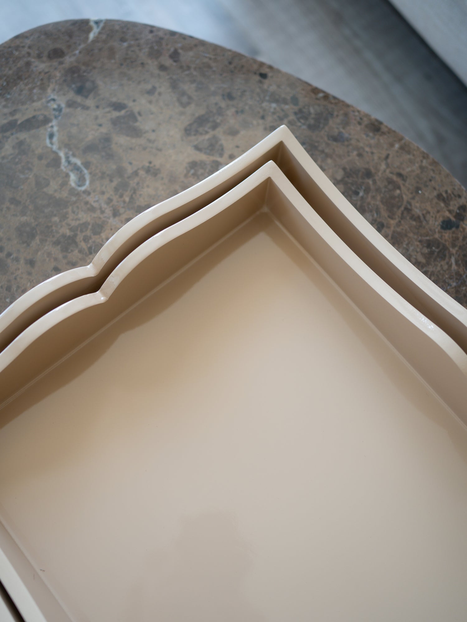 A close-up, high-angle view of a beige ceramic tray with a decorative wavy scalloped rim, nested inside a larger matching tray, set on a dark brown marble surface.