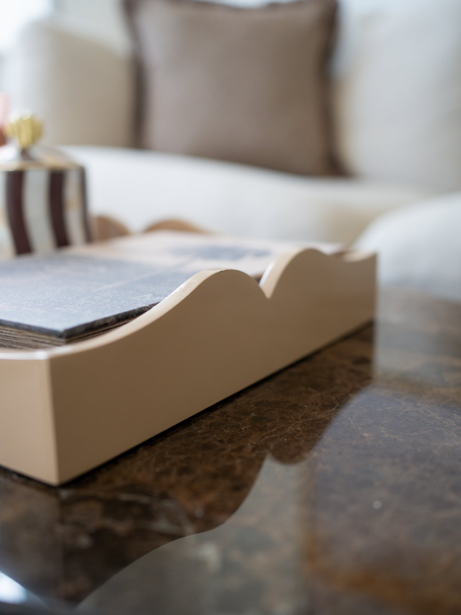 A close-up of a beige wooden tray with a scalloped edge, holding a stack of dark gray cards or booklets, set on a dark marble table with a blurred living room background.