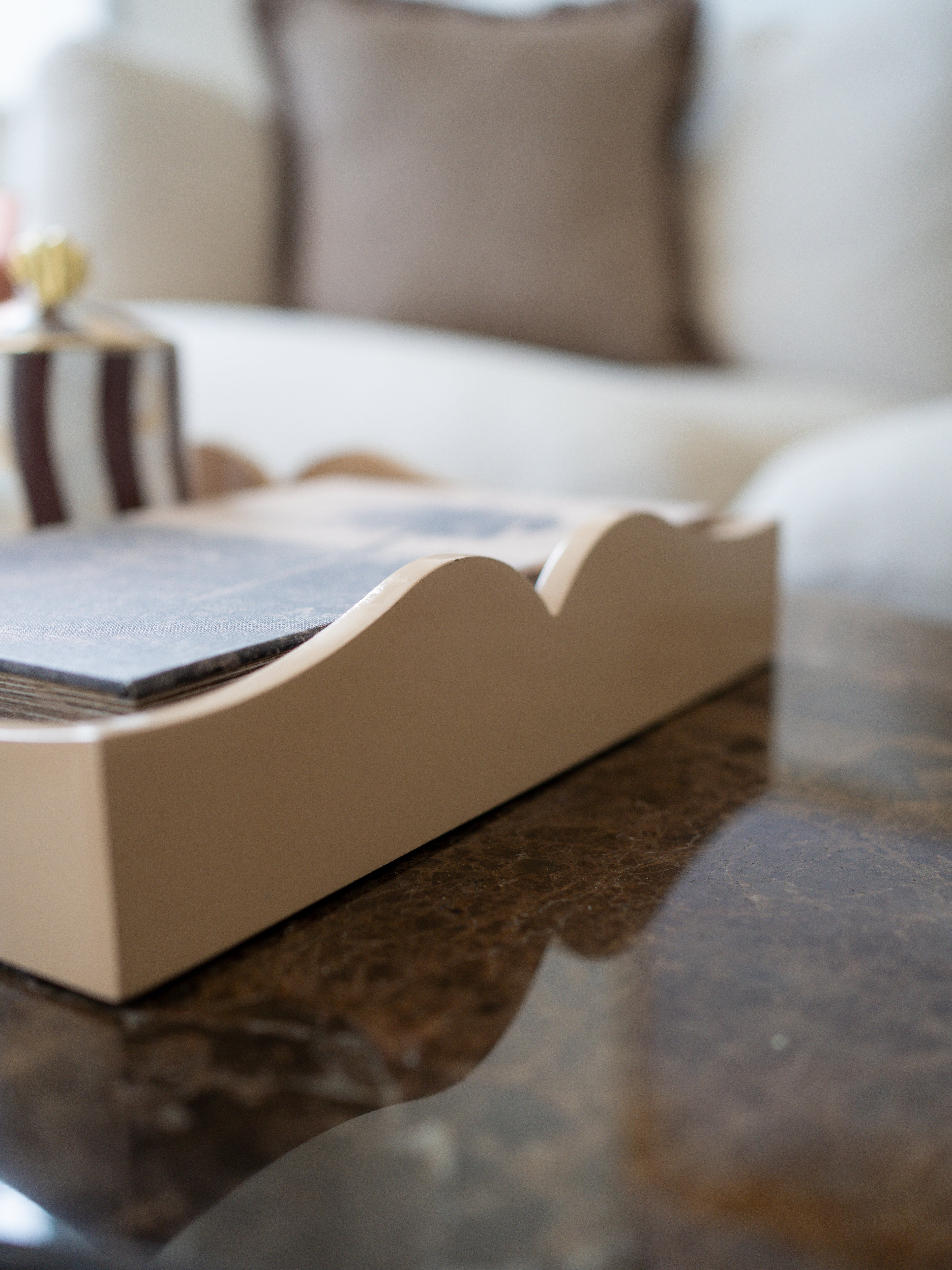 A close-up of a beige wooden tray with a scalloped edge, holding a stack of dark gray cards or booklets, set on a dark marble table with a blurred living room background.
