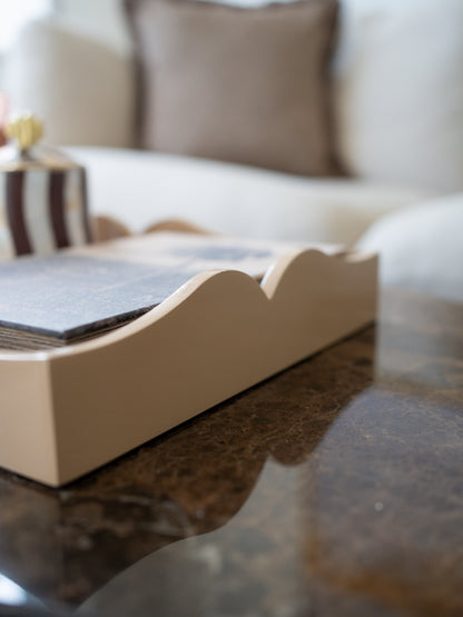 A close-up of a beige wooden tray with a scalloped edge, holding a stack of dark gray cards or booklets, set on a dark marble table with a blurred living room background.