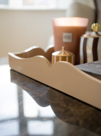 A close-up of a cream-colored wooden decorative tray with a wavy scalloped edge, resting on a dark marble tabletop with a softly blurred background featuring a candle and a striped container.