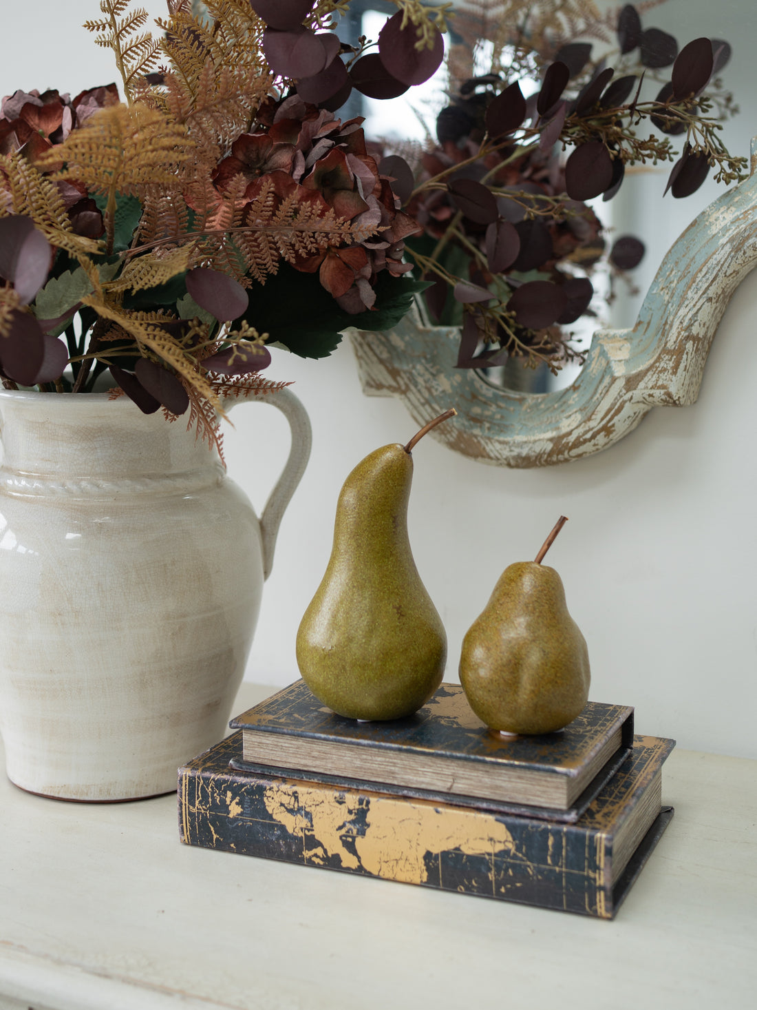 Two decorative pears placed on top of stacked vintage-style books on a white mantel, with a large cream pitcher of dried ferns and burgundy foliage beside them and a distressed mirror in the background.