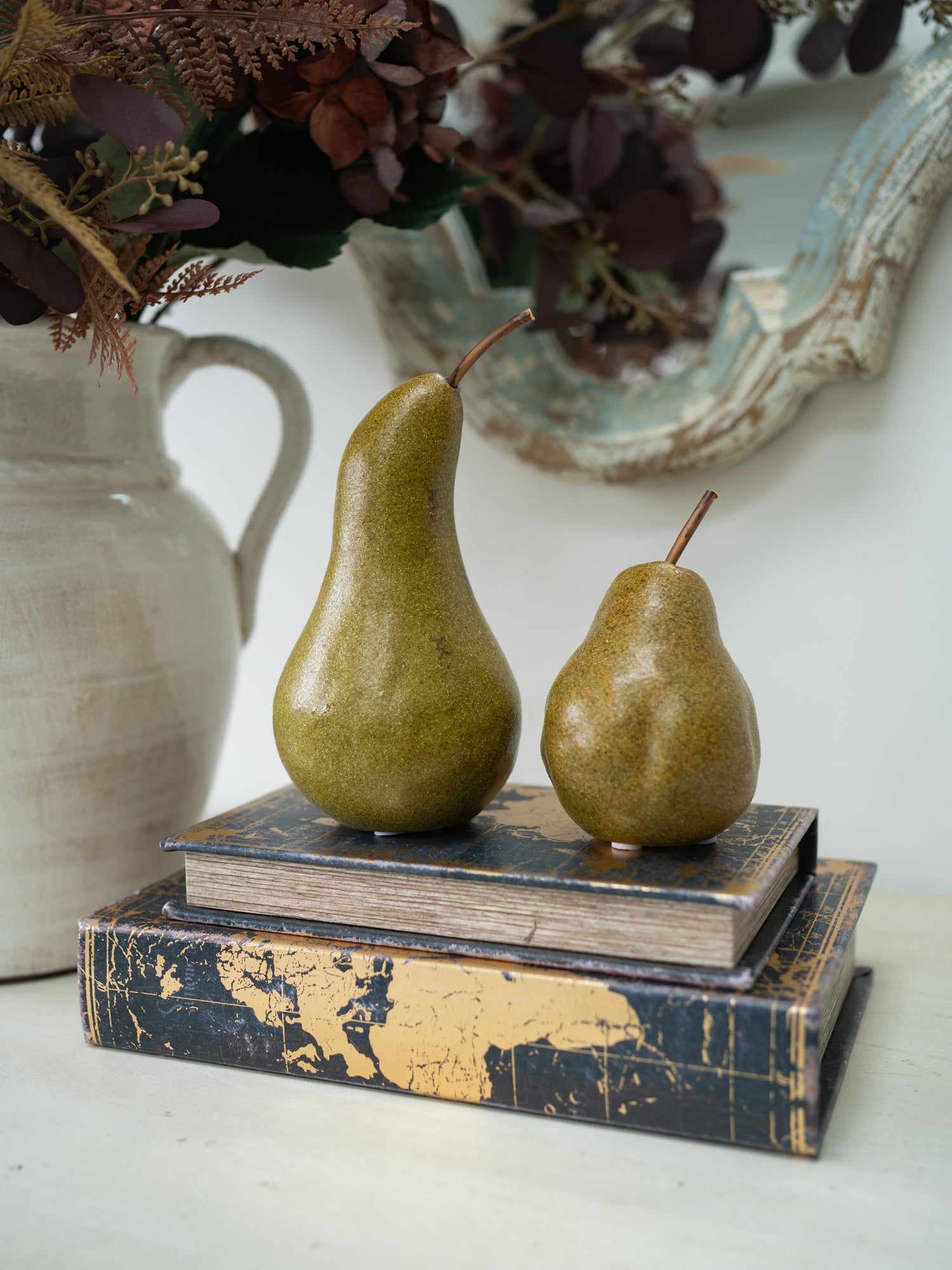 Two decorative pears with a rustic, distressed glaze sit on top of two vintage-style books. The books feature a dark navy blue and gold map design.
