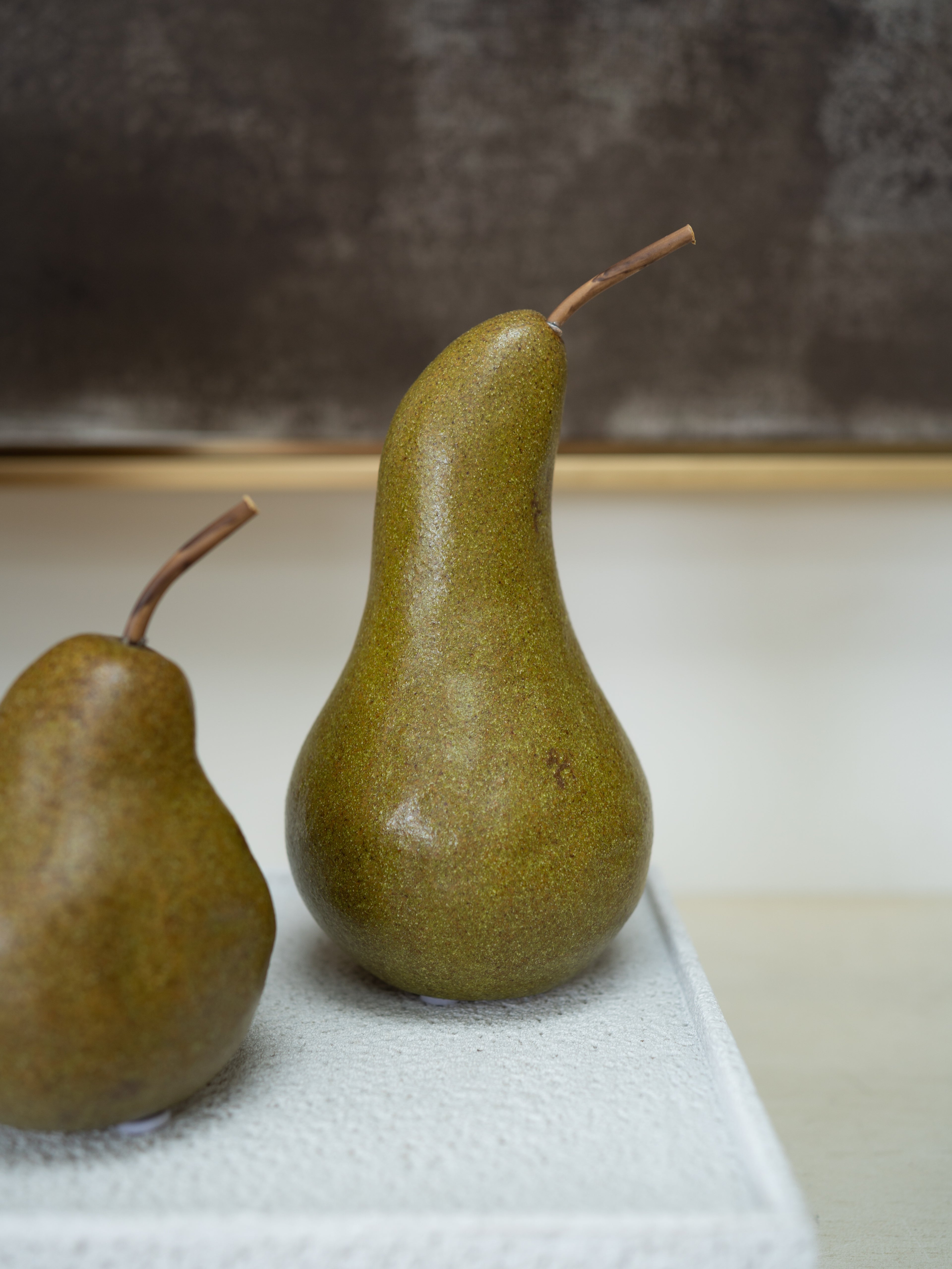 Two green pears with brown stems, one fully visible and one partially cropped on the left, resting on a textured white surface against a neutral gray and beige background.