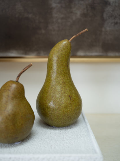 Two green pears with brown stems, one fully visible and one partially cropped on the left, resting on a textured white surface against a neutral gray and beige background.