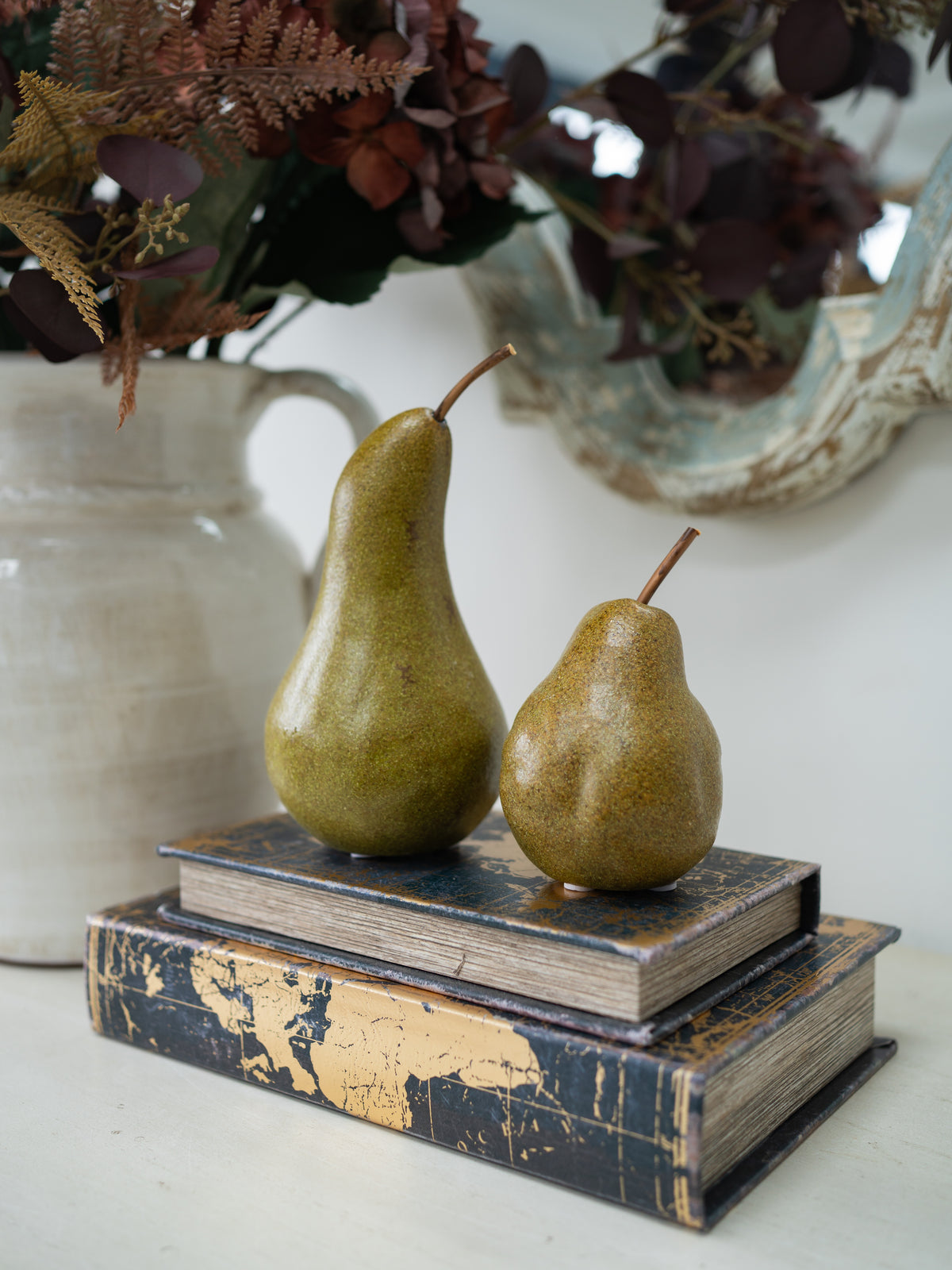 Two decorative pears placed on top of stacked vintage-style books, with a ceramic vase and dried floral arrangement blurred in the background.