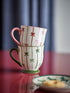 Two stacked ceramic mugs with white backgrounds, decorated with vertical red and green stripes and small bow motifs, set on a reflective red surface with a blurred curtain background.