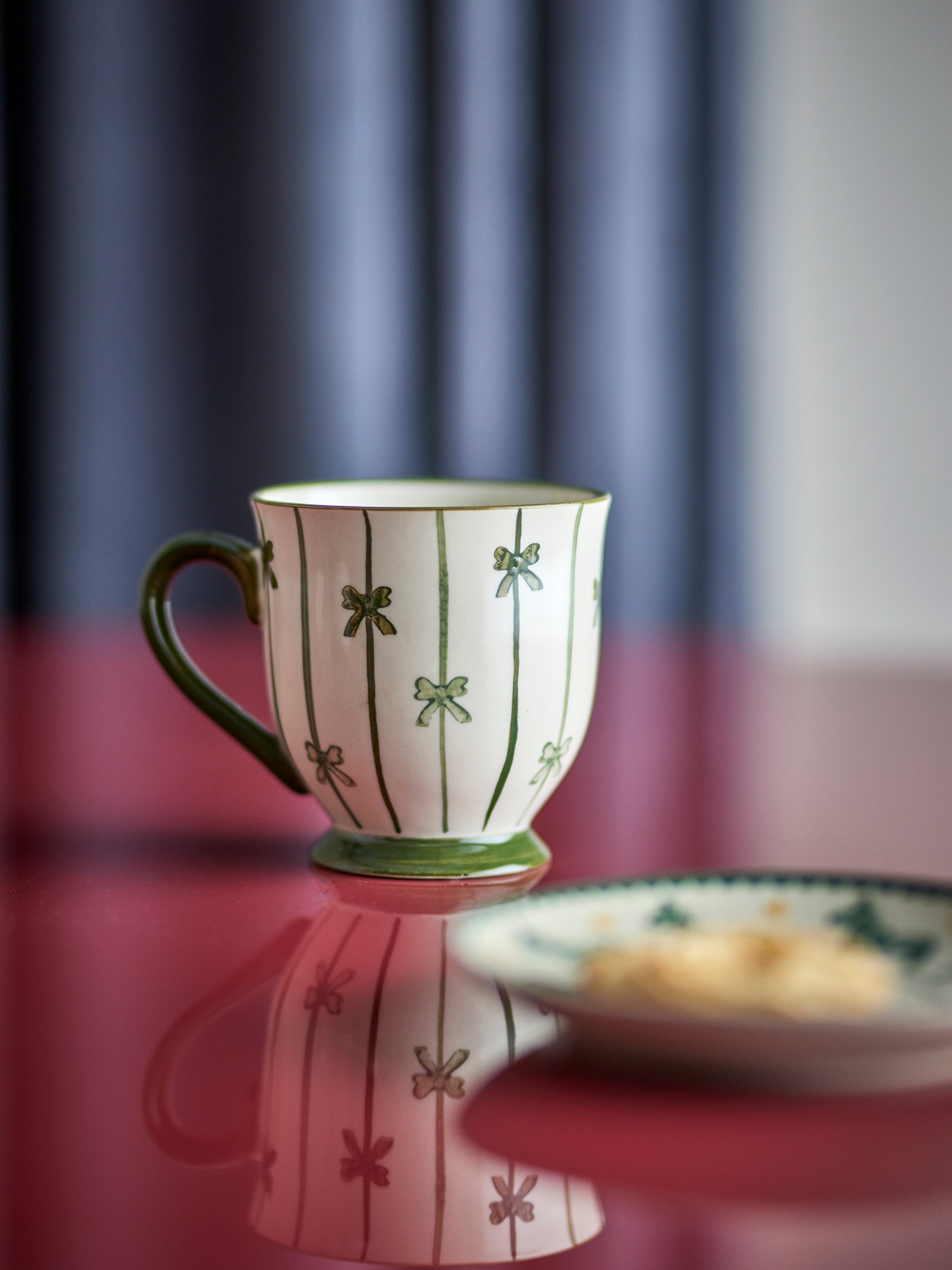 A white ceramic coffee mug with green vertical stripes and bow motifs, set on a reflective red surface next to a matching saucer with a piece of toast.