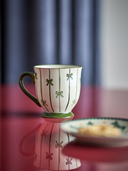 A white ceramic coffee mug with green vertical stripes and bow motifs, set on a reflective red surface next to a matching saucer with a piece of toast.