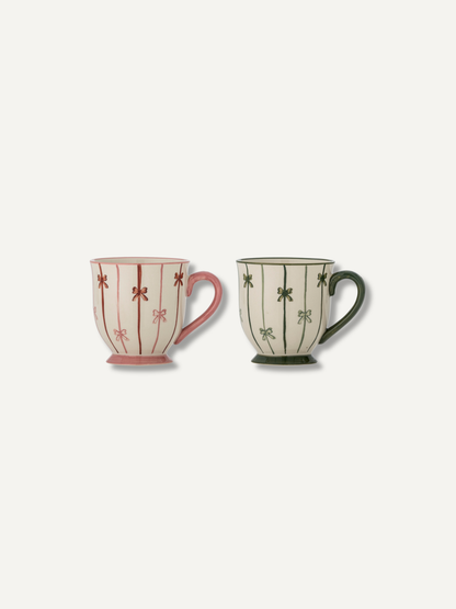 Two ceramic mugs with a vertical stripe pattern and small floral motifs, shown side by side on a light off-white background. The mug on the left is cream with pink stripes and handle, and the mug on the right is cream with dark green stripes and handle.