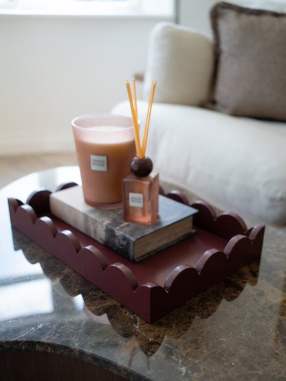 A scalloped oxblood wooden tray styled on a marble coffee table, holding a candle and a reed diffuser on top of a book, with a blurred living room background.