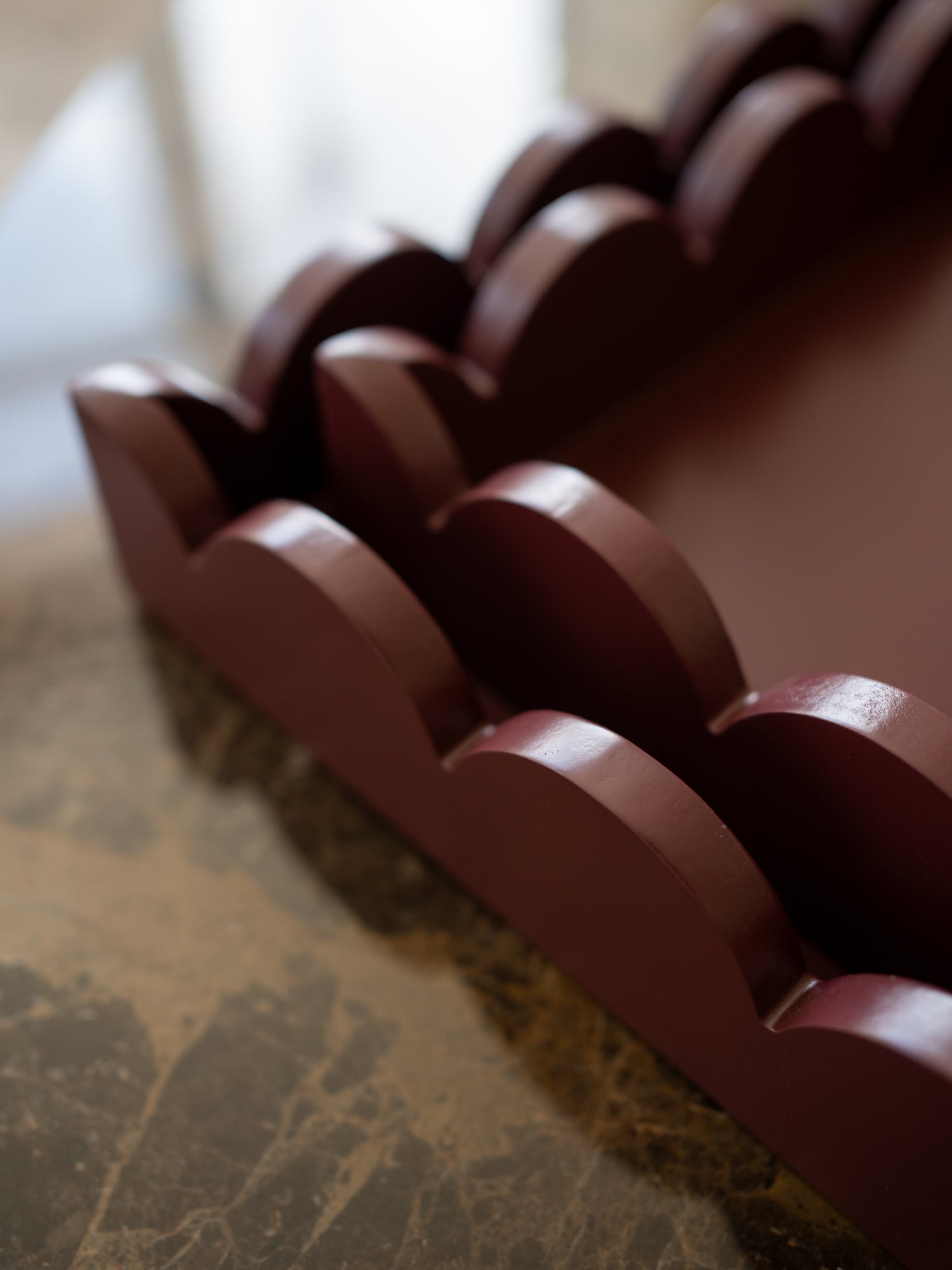 A close-up of a dark reddish-brown wooden tray or organizer with repeating scalloped or wave-like compartments, photographed on a marble-like surface with a softly blurred indoor background.
