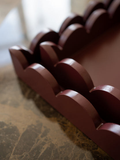 A close-up of a dark reddish-brown wooden tray or organizer with repeating scalloped or wave-like compartments, photographed on a marble-like surface with a softly blurred indoor background.