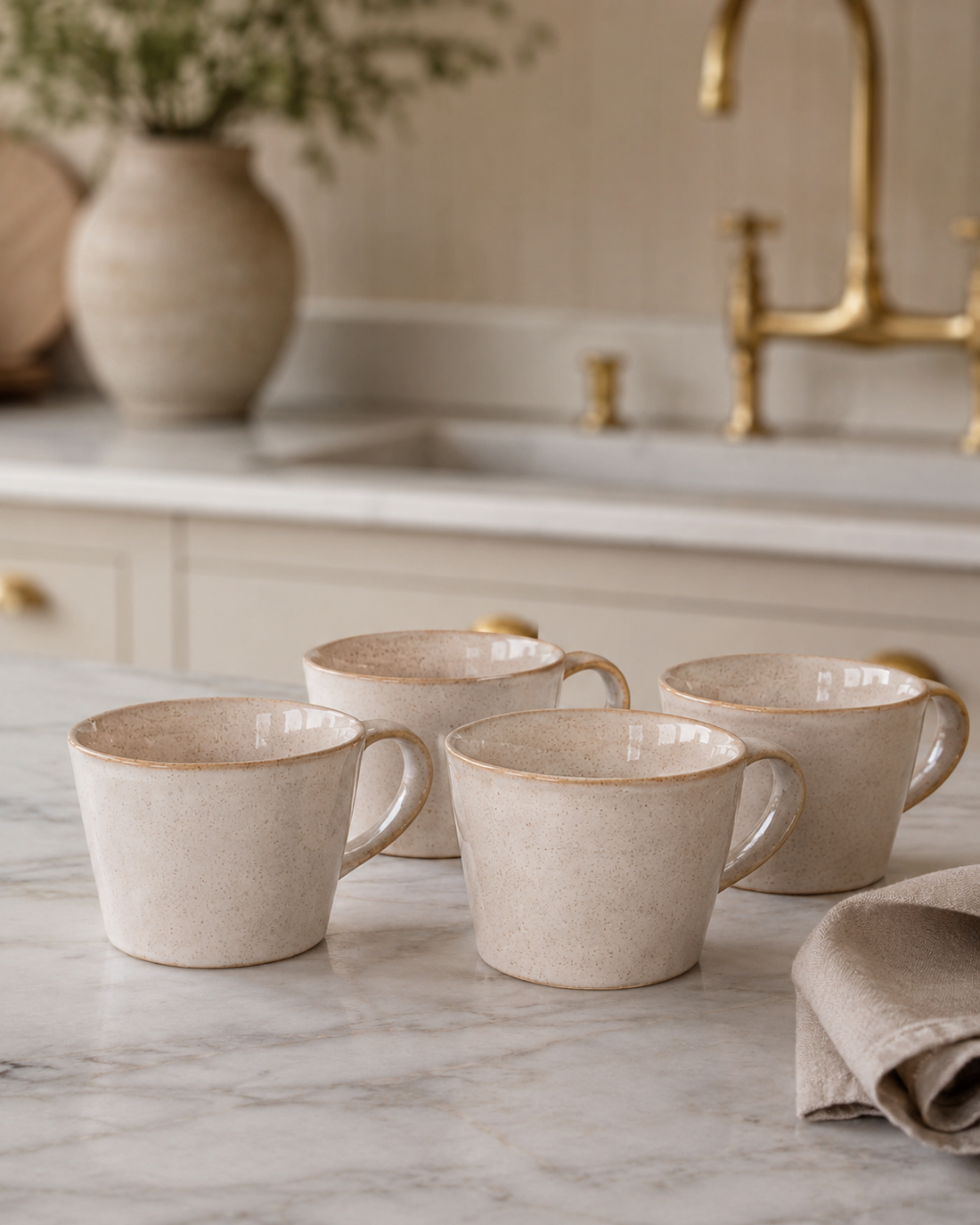 Set of four beige ceramic mugs on a marble countertop with a kitchen background.