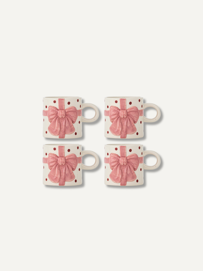 Four white ceramic mugs with pink gift bow and polka dot patterns, arranged in a 2x2 grid on a light off-white background.