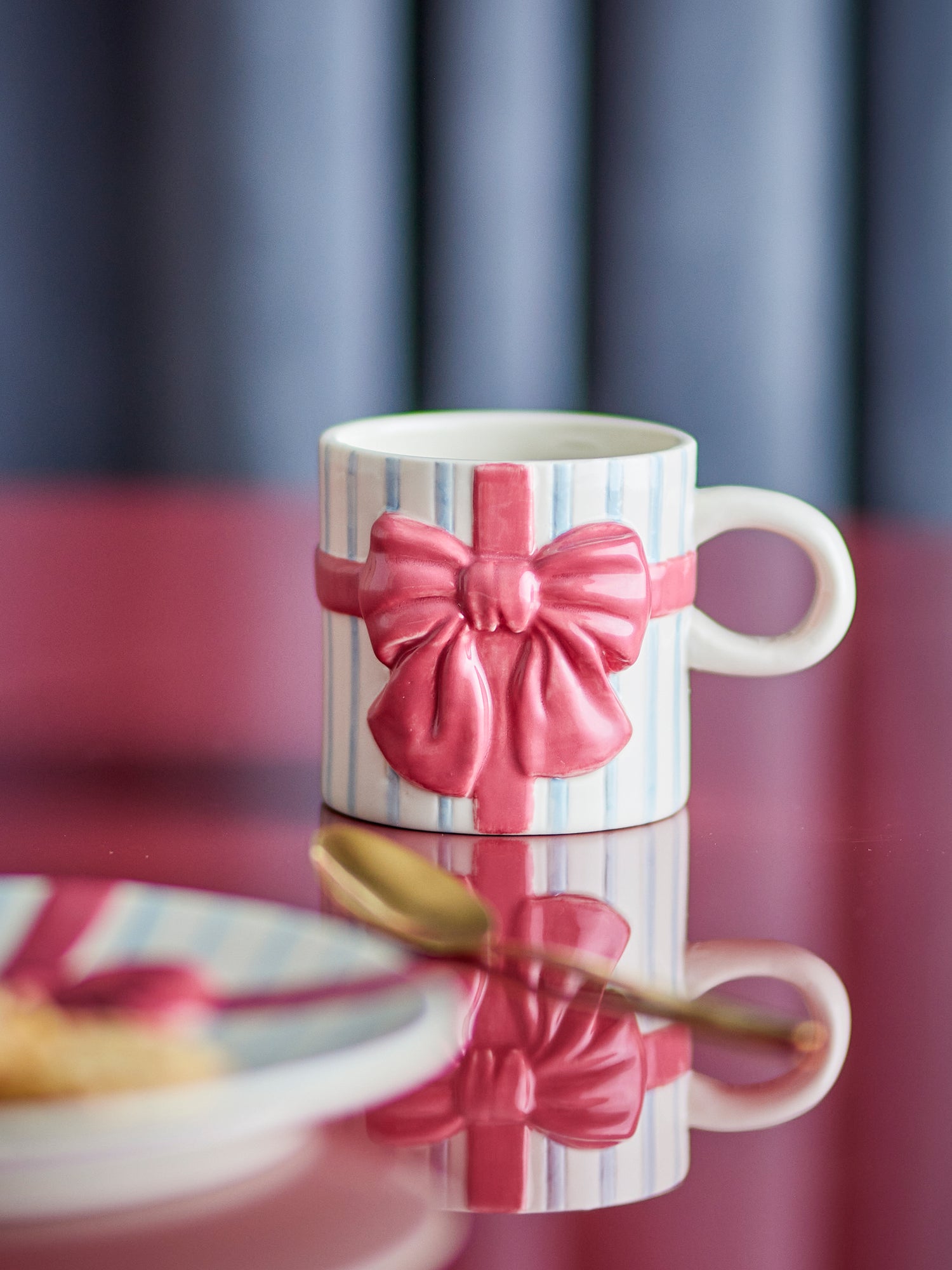 A white ceramic mug decorated with a pink 3D ribbon and bow design, set on a reflective dark surface with a blurred background.