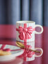 A white ceramic mug decorated with a pink 3D ribbon and bow design, set on a reflective dark surface with a blurred background.