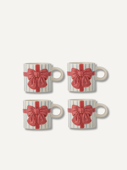 Four ceramic mugs arranged in a 2x2 grid, each decorated with a red gift bow and ribbon motif over a white and pale blue striped background, on a light off-white backdrop.